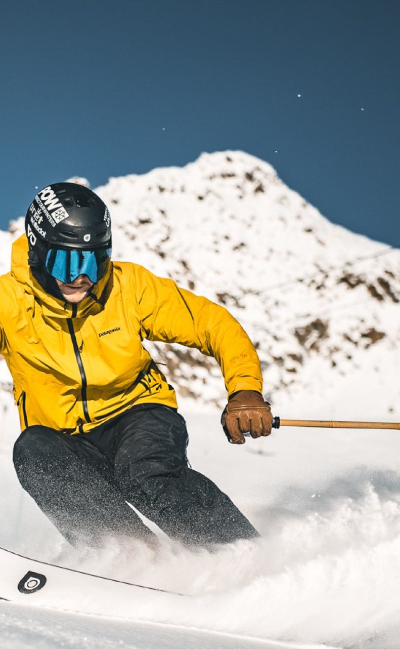 Skifahrer in gelber Jacke carvt bei blauem Himmel über die Piste, dahinter ein verschneiter Gipfel