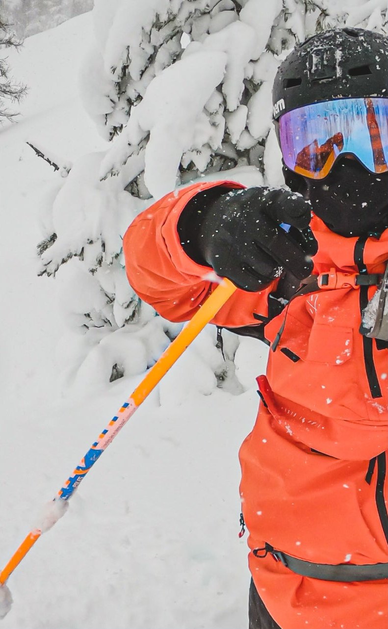 Skifahrer in oranger Jacke zeigt bei Schneefall zwischen verschneiten Bäumen in die Kamera